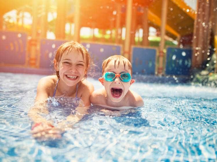 Little girl and her brother lying on front in shallow water in water park. The kids are laughing at camera, the boy is wearing swimming goggles.