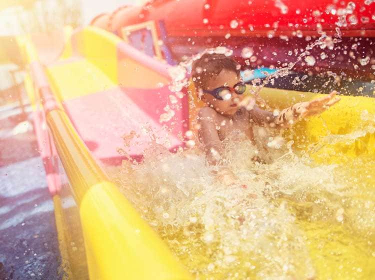 Little boy splashing into the water after sliding on water slide in water park. The boy aged 5 is wearing swimming goggles.