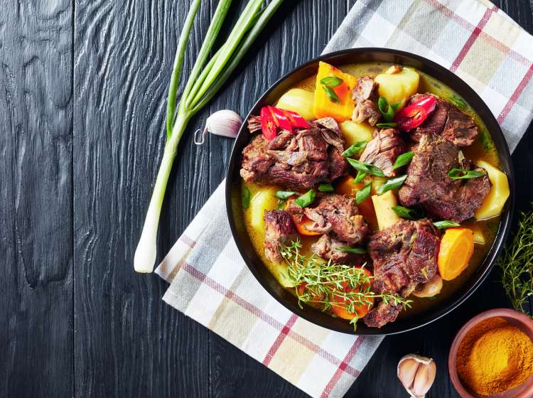 traditional Jamaican Curry Goat "u2013 slow cooked Jamaican Spiced meat and vegetables spicy Curry in a black bowl on a rustic wooden table, close-up