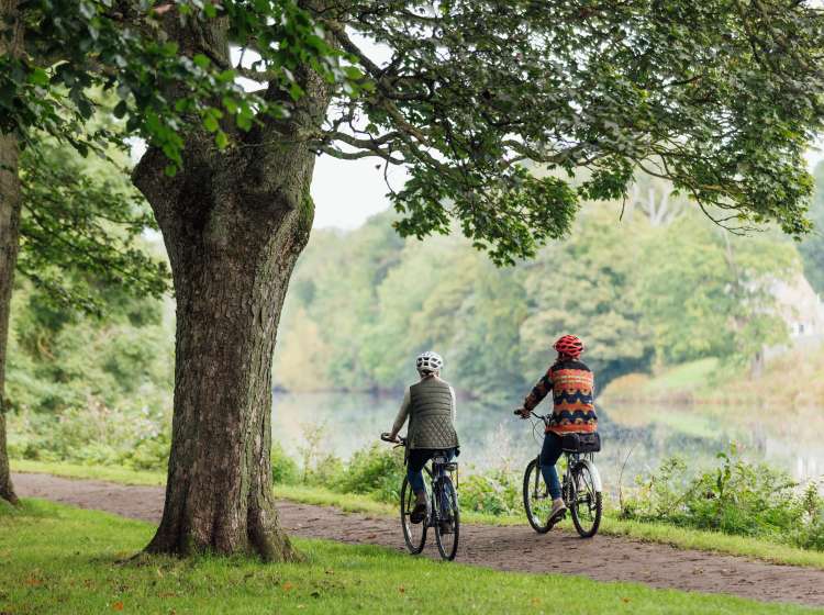 Rear view shot of an LGBTQI+ lesbian couples a bike ride together in a public park in Hexham, North East England. They are cycling side by side along a rivers edge.