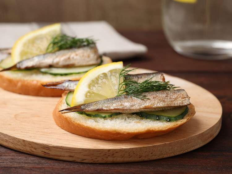 Delicious sandwich with sprats, cucumbers, lemon and dill on wooden board, closeup