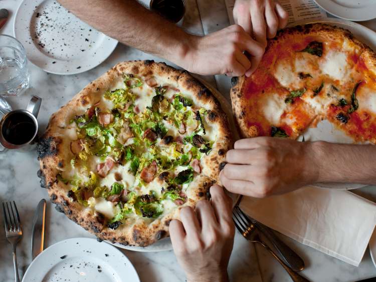Hands dig in to two pizzas (1 margherita, 1 with brussels sprouts and pancetta) on a marble table.