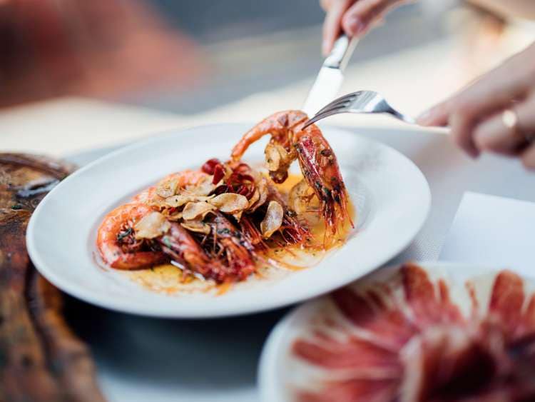 A plate of Spanish-style grilled shrimps, cooked with lemon juice, olive oil and sea salt. With Jamon Iberico in the background. Garlic prawn on a fork.