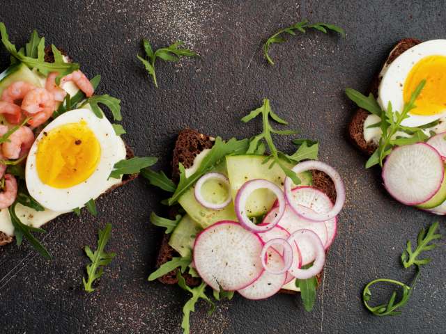 Open sandwiches on dark rye bread with eggs, shrimps, radishes, cucumber, cream cheese and arugula for breakfast on old dark background. Smorrebrod Traditional dish of Danish cuisine. Top view.; Shutterstock ID 2070570161; project: x; job: y; client: ; other: 