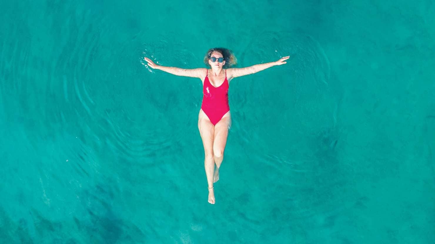 Aerial View of a Woman in Red Swimsuit Floating Serenely on the Crystal Clear Ocean Waters During Midday; Shutterstock ID 2443498231; purchase_order: -; job: -; client: -; other: