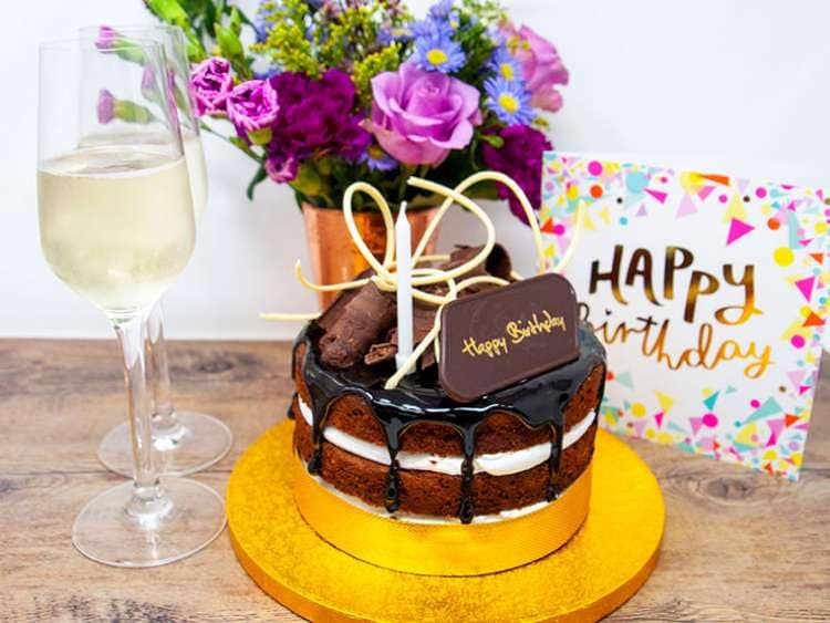 Cake with 'happy birthday' on surrounded by a couple of glasses of wine, flowers in a vase and a card