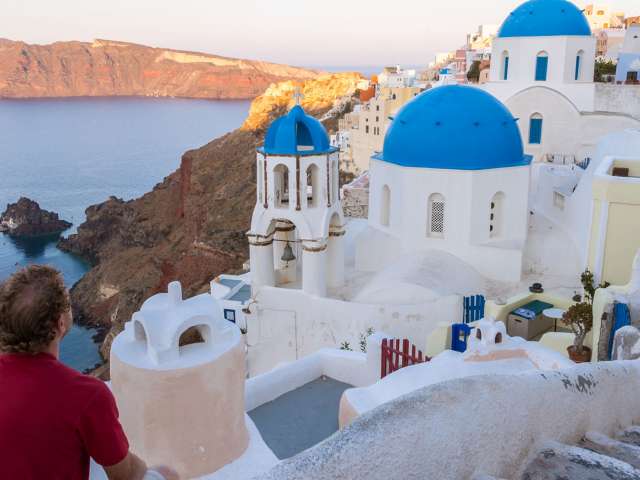 Tourist sitting on a wall and looking at town of Oia at sunrise, Santorini, Greece.