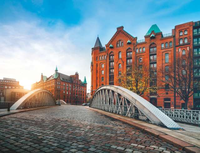 Arch bridge over alster canals with cobbled road in historical Speicherstadt of Hamburg, Germany, Europe. Scenic view of red brick building lit by golden sunset light.