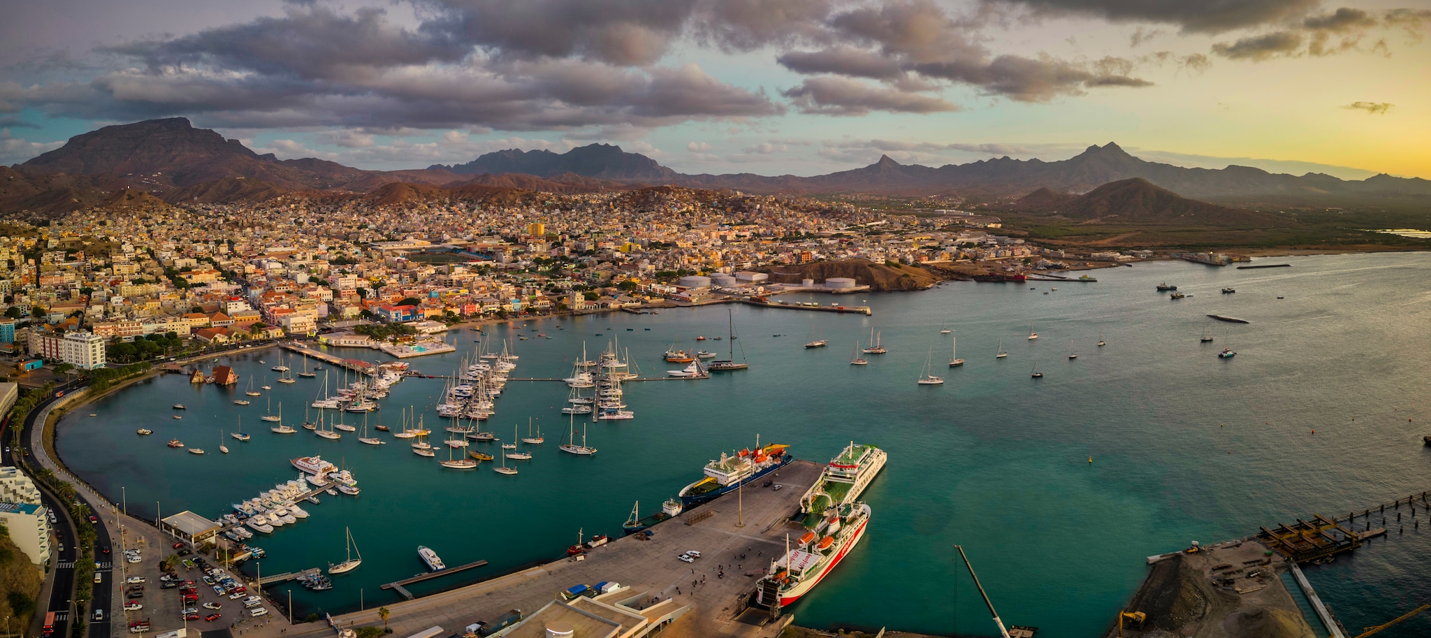{"metadata_en_GB":{"alt-text":"Panoramic aerial view of Mindelo city at sunset, with the marina and boats in the foreground, surrounded by the vibrant cityscape and mountains in the background under a warm, glowing sky"}}