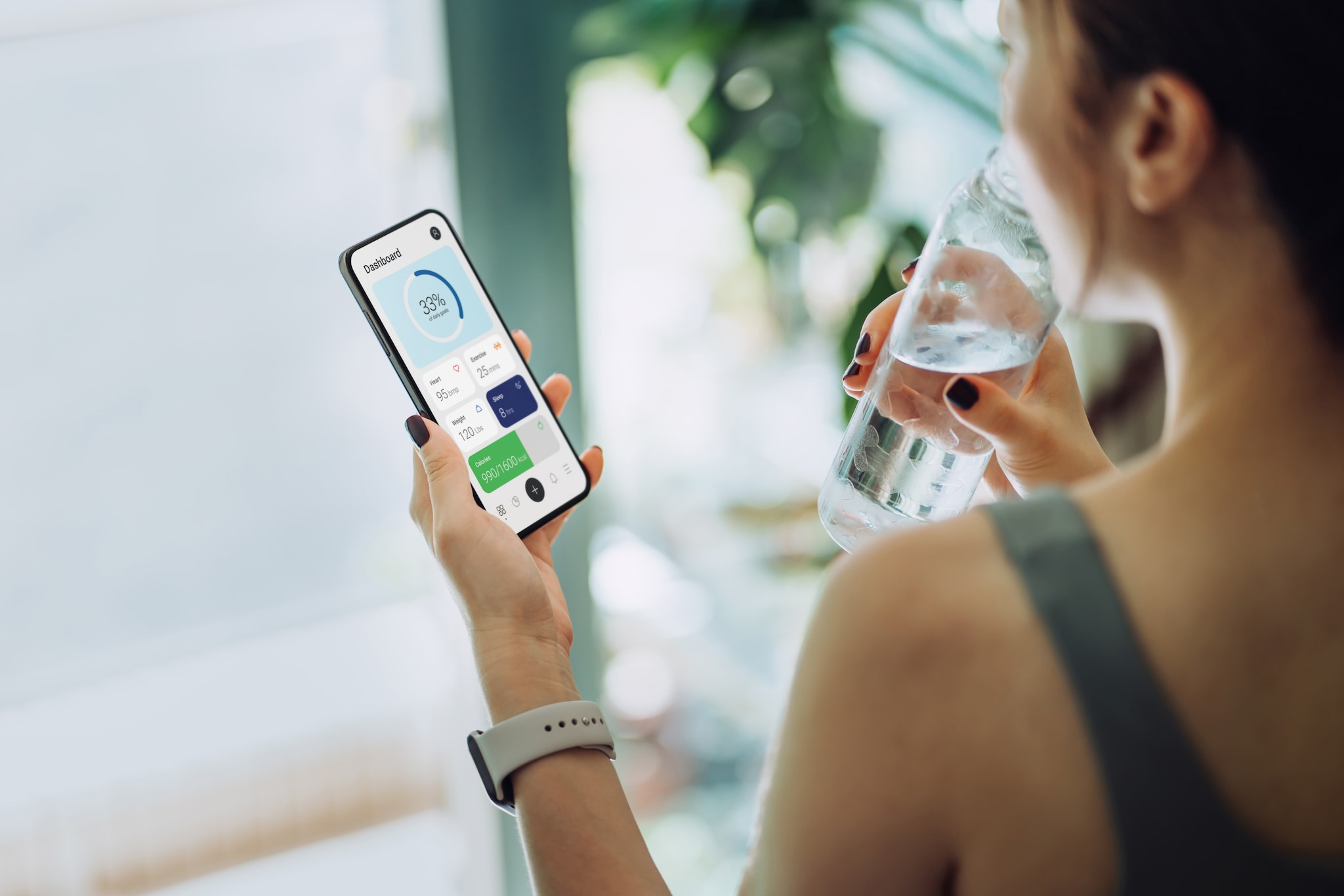 Over the shoulder view of young Asian sports woman refreshing with water and using fitness app on smartphone to monitor her training progress after fitness work out / exercising / practicing yoga at home in the fresh bright morning
