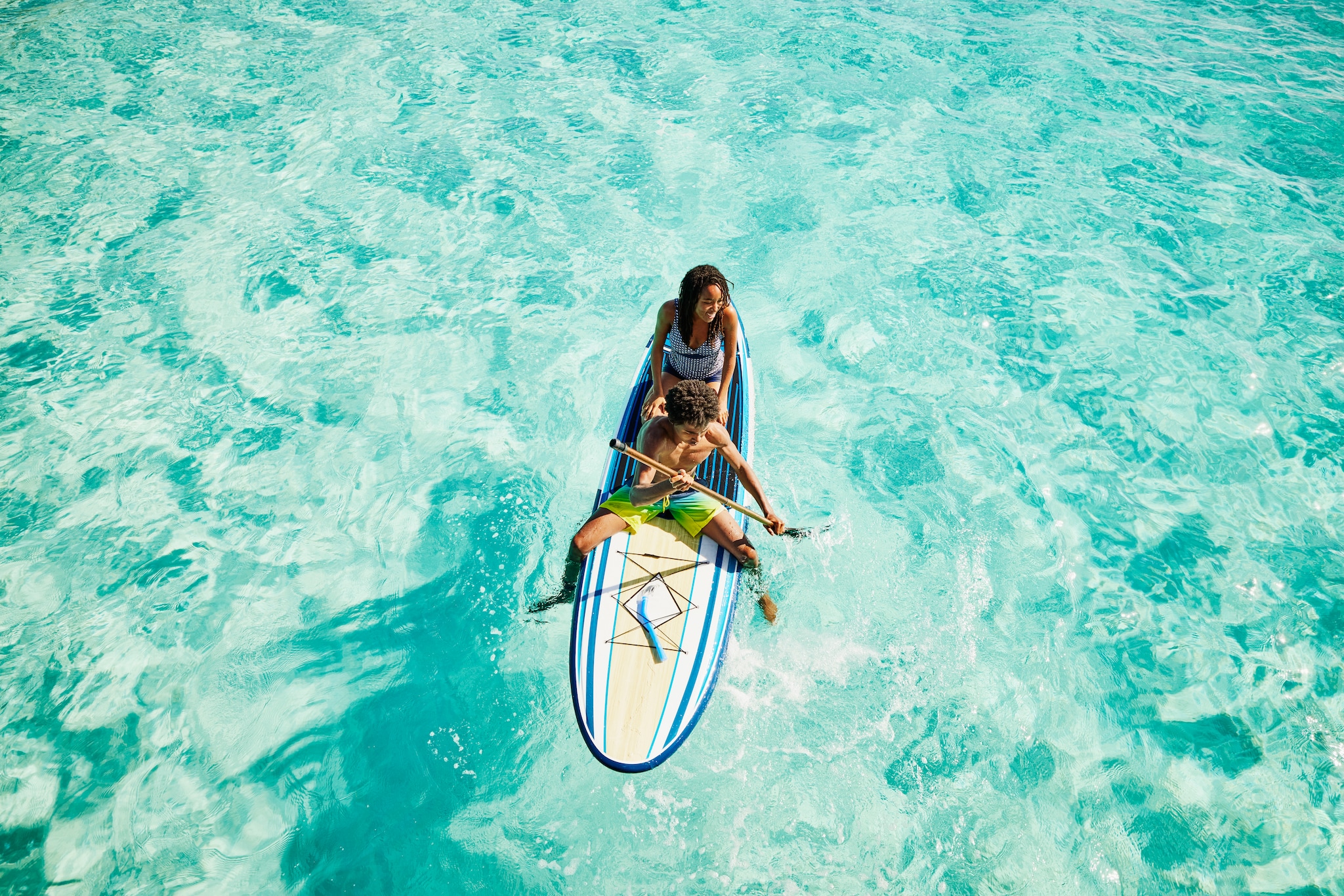 Wide shot of teenage brother and sister paddling paddleboard in ocean during family vacation in Caribbean Sea
